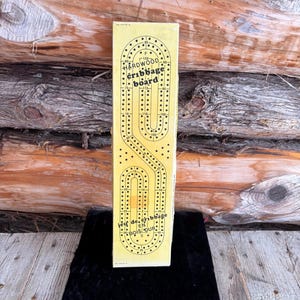 Könnte beinhalten: Eine gelbe Hartholz-Cribbage-Spielbox mit schwarzen Punkten und Text. Auf der Box steht "HARDWOOD cribbage board" und "Jeu de cribbage en bois dur". Das Spielbrett ist ein klassisches Brettspiel.