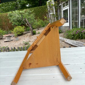May include: A light brown wooden tabletop easel with a triangular side profile, featuring a flat slanted surface for holding items, a small horizontal ledge, and four sturdy legs that provide a stable base for display purposes.