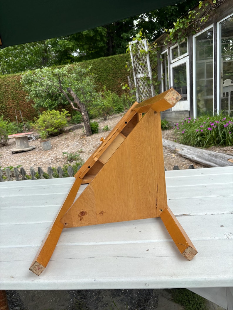 May include: A light brown wooden tabletop easel with a triangular side profile, featuring a flat slanted surface for holding items, a small horizontal ledge, and four sturdy legs that provide a stable base for display purposes.