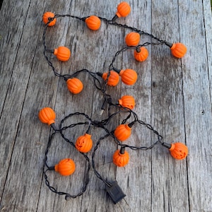 May include: A string of decorative lights featuring small, orange, pumpkin-shaped covers with carved jack-o'-lantern faces. The lights are connected by a thin black electrical cord that includes a standard two-prong plug at the end of the strand.