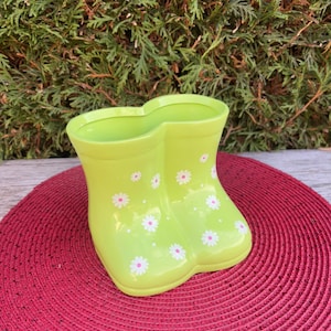 Peut inclure: Un pot de fleurs en céramique vert citron en forme de bottes de pluie, décoré de marguerites blanches. Le pot est posé sur un tapis rouge tressé, avec un fond de feuillage vert.