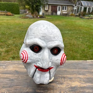 May include: A white mask with red and white spiral cheeks, red lips, and black eyes. The mask has a textured surface, suitable for a Halloween costume or horror-themed event.