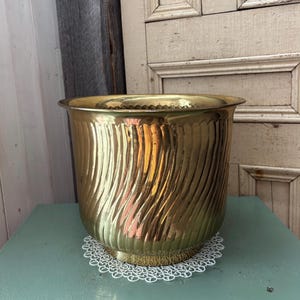 May include: A large, gold-coloured metal planter with a swirling, textured design. The planter has a wide, slightly flared rim and sits on a decorative white lace doily. The planter is on a light green surface.