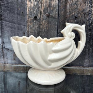 Peut inclure: Un pot en céramique blanc en forme de coquille avec un motif cannelé et une poignée. Le pot a une base et est parfait pour présenter des plantes ou des fleurs.