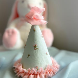 May include: A light green party hat with a pink ruffled trim and a pink fabric flower on top. The hat is embroidered with small pink flowers.
