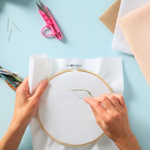 May include: A person is working on a cross stitch project. They are using a wooden embroidery hoop with white Aida cloth stretched over it. A green thread is being threaded through the fabric with a needle.