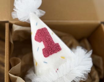 May include: White felt party hat with a pink felt number one embroidered on the front. The hat has a white tulle ruffle around the bottom and a white tulle bow on top. The hat is decorated with colorful sprinkles.