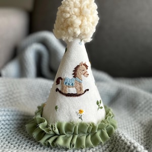 May include: A white fabric party hat with a green ruffle trim. The hat is decorated with an embroidered rocking horse and small flowers.