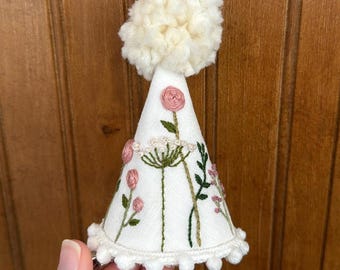 May include: A white party hat with a white pom-pom on top. The hat is decorated with pink and green embroidered flowers.