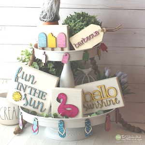 May include: A tiered tray decorated with summer-themed wooden signs. The top tier features a gnome wearing sunglasses, popsicle signs, and a sign that says "barbecue". The middle tier has a sign that says "fun in the sun", a flamingo float, and a sign that says "hello sunshine". The bottom tier has a sign that says "cream" and flip-flop shaped signs.