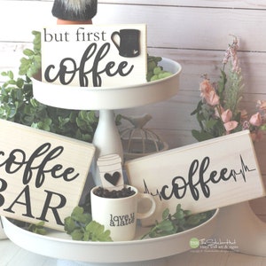 May include: A tiered tray with three wooden signs that say "but first coffee", "coffee bar", and "coffee" with a heart beat graphic. The tray is decorated with a gnome, coffee beans, a coffee cup, and greenery.