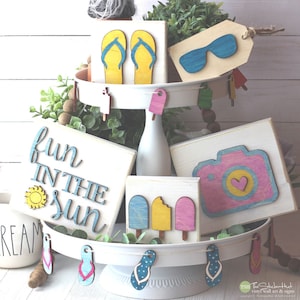 May include: A tiered tray decorated with summer-themed wooden signs. The signs feature flip flops, sunglasses, ice cream, a camera, and the text "Fun in the Sun".