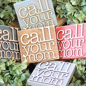 May include: A collection of square wooden signs in various colors, including blue, brown, red, and gray, each displaying the phrase "Call Your Mom" in a layered, three-dimensional design. The signs are arranged on a bed of green foliage.