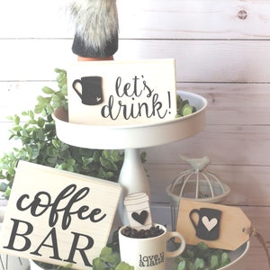May include: A tiered white cake stand with a coffee theme. The top tier has a wooden sign that says "let's drink!" with a black coffee cup graphic. The bottom tier has a wooden sign that says "coffee BAR" and a white coffee mug with the text "love u a latte" and black coffee beans.