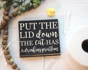 Puede incluir: Un letrero cuadrado de madera negra con bordes desgastados y texto en blanco que dice: PUT THE LID down THE cat HAS A drinking problem. El letrero se muestra junto a un collar de cuentas de madera, un pato de goma amarillo y bastoncillos.