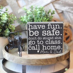May include: A black wooden sign with white text that reads "have fun. be safe. don't do stupid shit. go to class. call home. Mom & Dad." The sign is on a wooden tray with a small decorative bicycle.