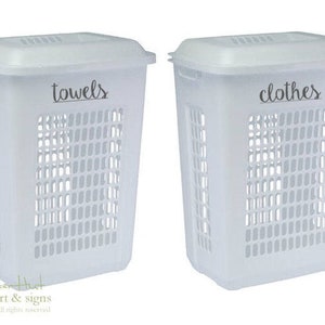 May include: Two white plastic laundry hampers with lids. The hamper on the left has the word "towels" printed on the front. The hamper on the right has the word "clothes" printed on the front.