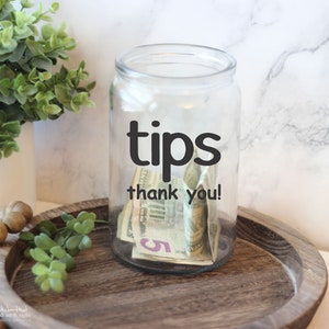May include: A clear glass jar with a black vinyl sticker that says "tips thank you!"  The jar is filled with US dollar bills.