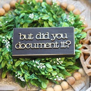 May include: A black wooden sign with gold lettering that reads "but did you document it?" The sign is sitting on a bed of green foliage.
