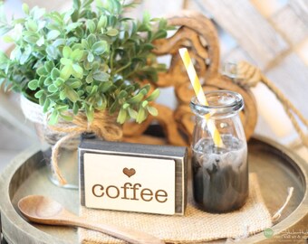 May include: A small rectangular wooden sign with a white front panel featuring a brown heart icon and the text "coffee" in lowercase letters. Next to it is a clear glass bottle filled with a dark beverage, a yellow and white striped straw, and a wooden spoon.