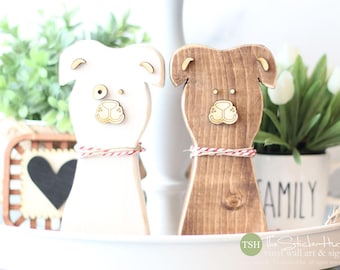 May include: Two wooden dog figurines, one white and one brown, with painted details and a red and white twine bow around their necks. The figurines are sitting on a white tiered tray.