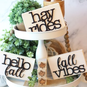 May include: A white tiered tray with fall-themed wooden signs. The signs read "hay rides", "hello fall", and "fall vibes". There are also three small wooden blocks with a leaf design.