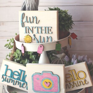 May include: A tiered tray decorated with summer-themed wooden signs. The top tier features a sign that says "fun in the sun" with a yellow sun cutout. The middle tier has three signs: "hello summer", a camera with a pink and yellow design, and "hello sunshine". The bottom tier has a white base with a string of flip-flop cutouts.