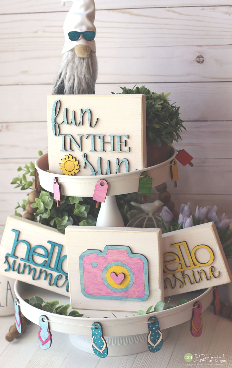 May include: A tiered tray decorated with summer-themed wooden signs. The top tier features a sign that says "fun in the sun" with a yellow sun cutout. The middle tier has three signs: "hello summer", a camera with a pink and yellow design, and "hello sunshine". The bottom tier has a white base with a string of flip-flop cutouts.