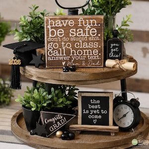 May include: A two-tiered wooden tray with graduation-themed decorations. Includes a miniature graduation cap, signs with inspirational quotes, a small clock, and decorative plants. The signs read "have fun. be safe," and "the best is yet to come."