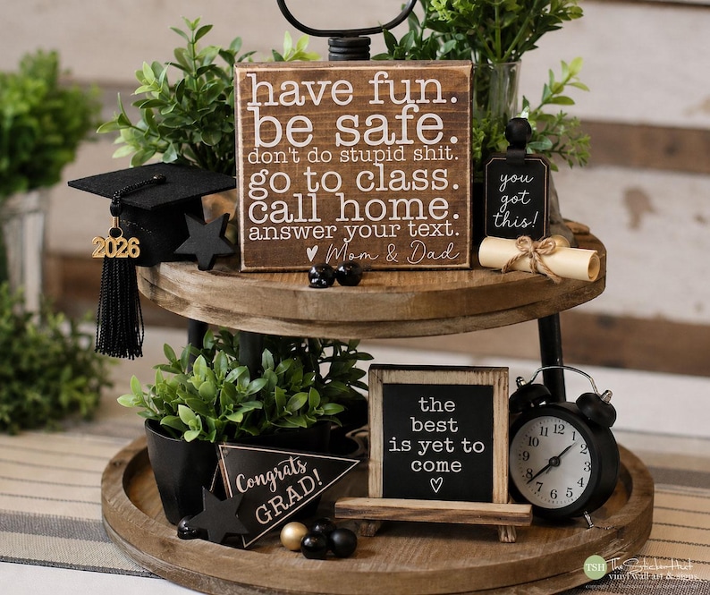 May include: A two-tiered wooden tray with graduation-themed decorations. Includes a miniature graduation cap, signs with inspirational quotes, a small clock, and decorative plants. The signs read "have fun. be safe," and "the best is yet to come."
