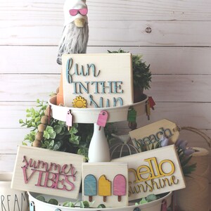 May include: A tiered tray decorated with summer-themed wooden signs, including "fun in the sun", "summer vibes", "hello sunshine", and "beach". The tray is also decorated with wooden popsicle and flip-flop shapes.