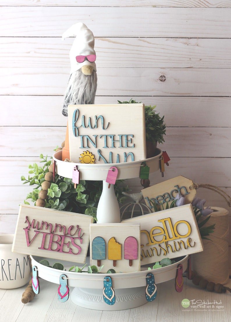 May include: A tiered tray decorated with summer-themed wooden signs, including "fun in the sun", "summer vibes", "hello sunshine", and "beach". The tray is also decorated with wooden popsicle and flip-flop shapes.