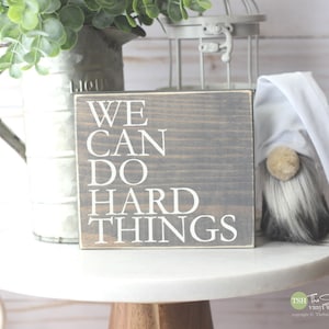 May include: A gray wooden sign with white text that reads "WE CAN DO HARD THINGS". The sign is sitting on a white table with a small gray and white gnome figurine to the right.
