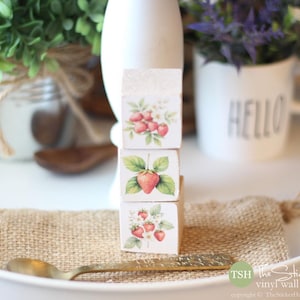 May include: Three stacked wooden blocks with strawberry illustrations. Each block features a different strawberry design with red berries and green leaves. The blocks sit on a burlap cloth with a gold spoon.