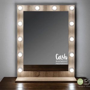 May include: A wooden vanity mirror with a light wood finish and a rectangular mirror. The mirror has a border of light bulbs around the perimeter. The mirror is sitting on a wooden table. The text "Cash Payment Preferred" is printed on the mirror.
