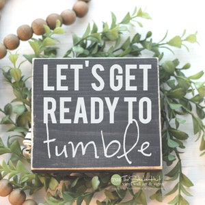May include: A black wooden sign with white text that reads "Let's Get Ready To Tumble". The sign is sitting on a white surface with green plants and brown wooden beads.