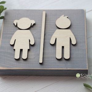 May include: A gray wooden sign with a cutout of a girl and a boy on either side of a vertical line. The sign is distressed and has a rustic look.