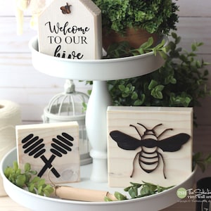 May include: A white tiered tray with three wooden signs. The top sign says "Welcome to our hive". The middle sign has a black silhouette of two honey dippers. The bottom sign has a black silhouette of a bee.