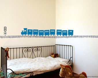 May include: A blue train engine and boxcars with black wheels on a black track with a traffic light on a white wall. The train is a wall decal.