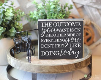 May include: A black and white wooden sign with the text "THE OUTCOME you WANT IS ON THE OTHER SIDE OF EVERYTHING you DON'T FEEL LIKE DOING today." The sign is on a wooden tray with a small decorative bicycle.