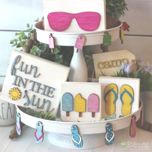 May include: A tiered tray decorated with summer-themed wooden signs. The top tier features a sign with pink sunglasses, the middle tier has a sign that says "fun in the sun" with a yellow sun, and the bottom tier has a sign that says "camp" and a sign with two blue flip flops. The tray is decorated with wooden cutouts of popsicles and flip flops.