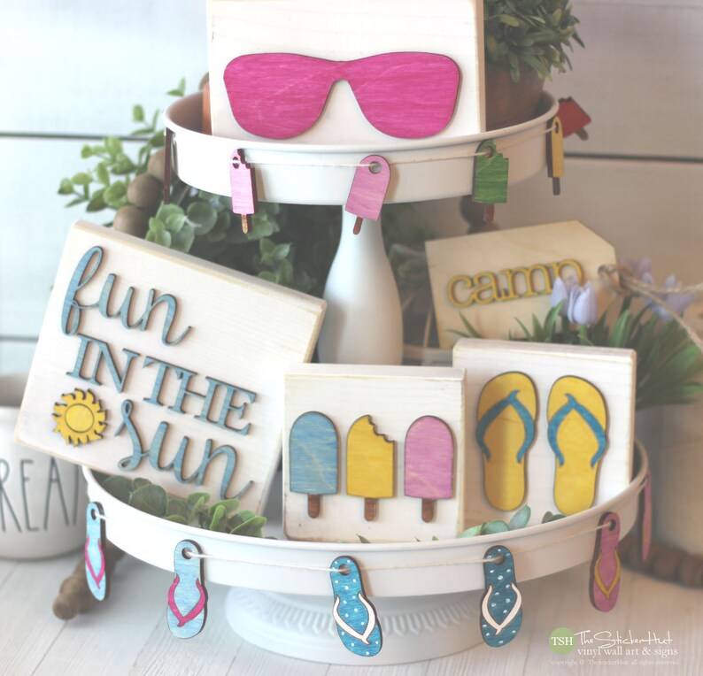 May include: A tiered tray decorated with summer-themed wooden signs. The top tier features a sign with pink sunglasses, the middle tier has a sign that says "fun in the sun" with a yellow sun, and the bottom tier has a sign that says "camp" and a sign with two blue flip flops. The tray is decorated with wooden cutouts of popsicles and flip flops.