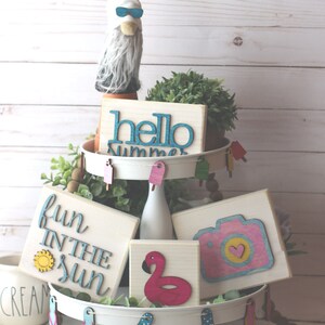 May include: A white tiered tray decorated with summer-themed wooden signs. The signs feature phrases like "hello summer", "fun in the sun", and a camera with a heart. The tray is also decorated with small wooden flip flops and popsicles.