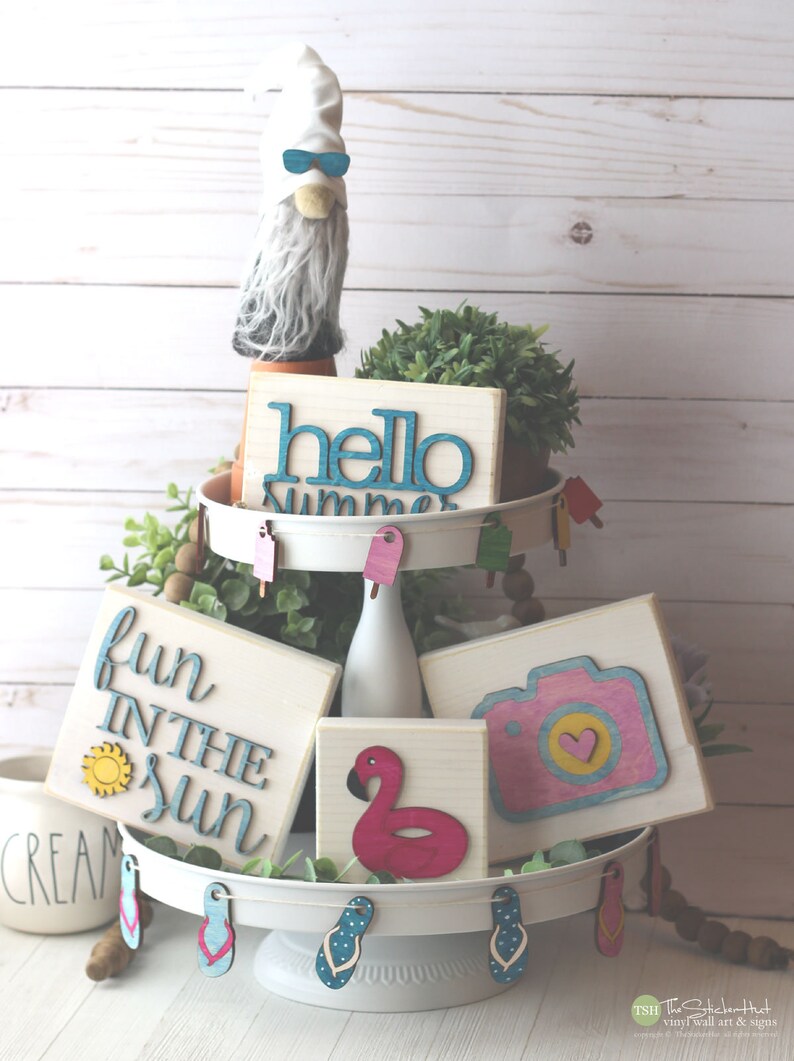 May include: A white tiered tray decorated with summer-themed wooden signs. The signs feature phrases like "hello summer", "fun in the sun", and a camera with a heart. The tray is also decorated with small wooden flip flops and popsicles.
