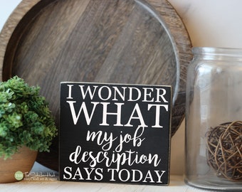 May include: Black wooden sign with white text that reads "I WONDER WHAT my job description SAYS TODAY".
