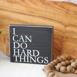 May include: A black wooden sign with white lettering that reads "I CAN DO HARD THINGS".