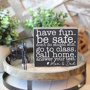 May include: A black wooden sign with white text that reads "have fun. be safe. don't do stupid shit. go to class. call home. answer your text. Mom & Dad." The sign is on a wooden tray with a small decorative bicycle.