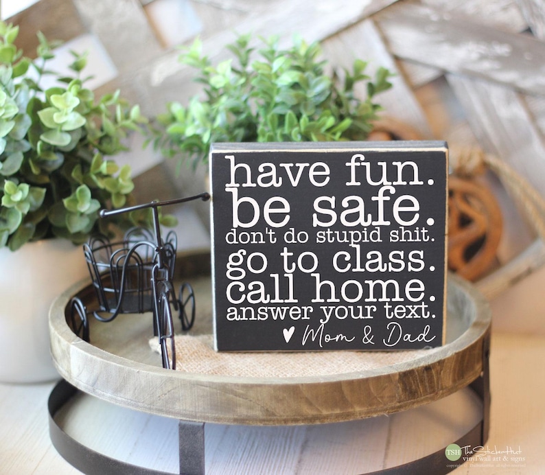 May include: A black wooden sign with white text that reads "have fun. be safe. don't do stupid shit. go to class. call home. answer your text. Mom & Dad." The sign is on a wooden tray with a small decorative bicycle.