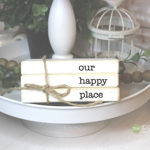 May include: A stack of three white wooden blocks with the words "our happy place" written in black. The blocks are tied together with twine and are sitting on a white cake stand.