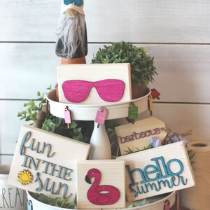May include: A tiered tray decorated with summer-themed wooden signs. The signs feature phrases like "fun in the sun", "hello summer", and "barbecue". There are also pink sunglasses, a pink flamingo, and flip-flops.